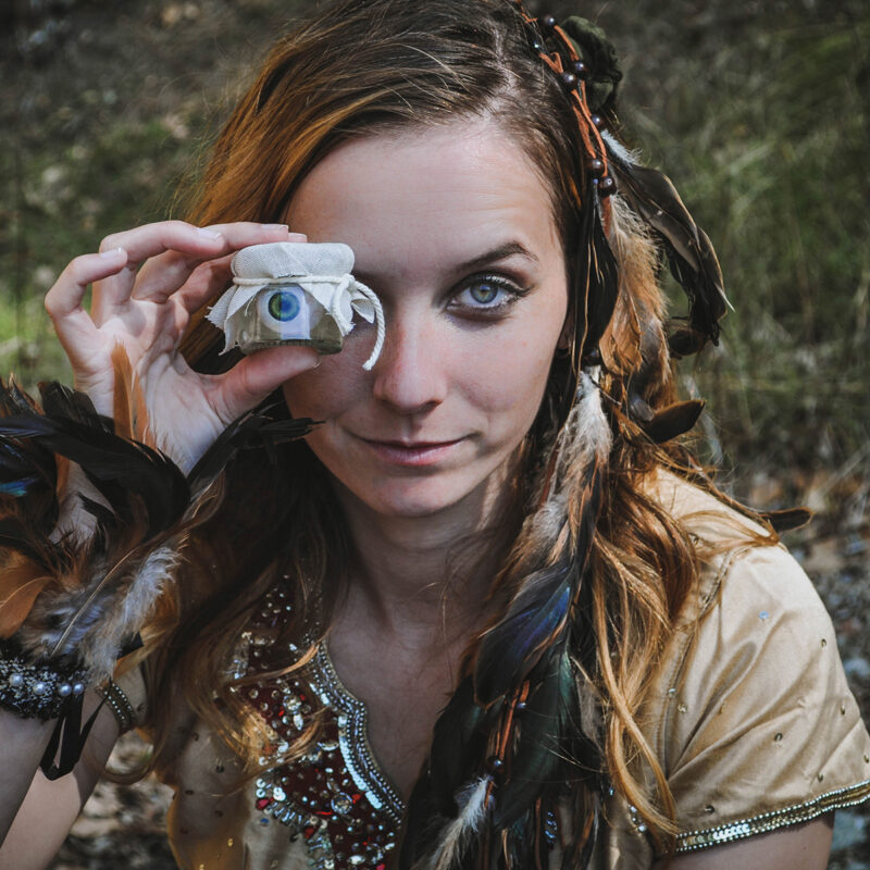 Femme pirate avec des plumes sur la tête, elle tient un bocal contenant un oeil devant son propre oeil